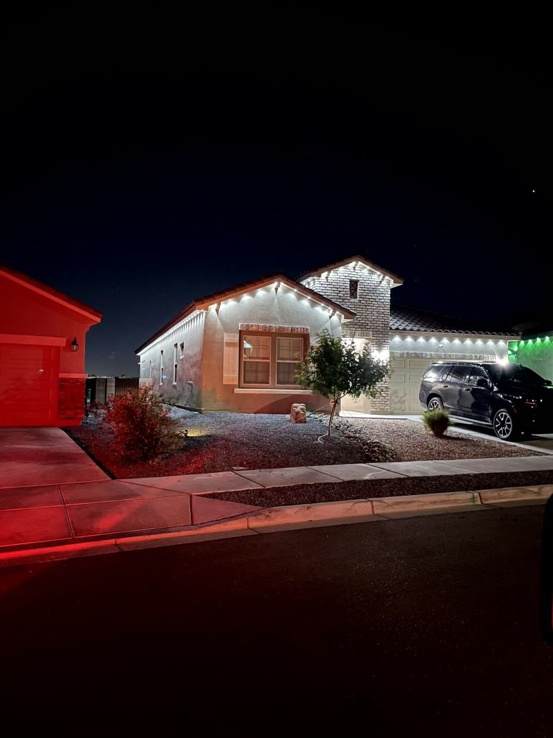 Warm white permanent lighting along a single-story Albuquerque roofline at night, with a neighboring elevation washed red